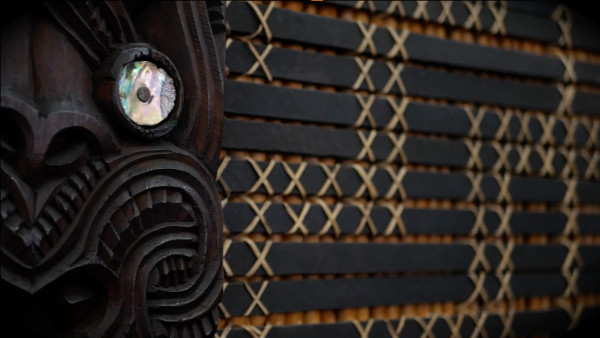 A wooden panel on a wall with intricate cross patterns made of reed, and a carved wooden face with paua shell eyes.