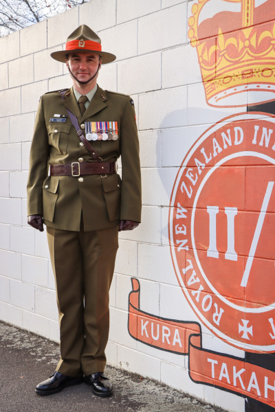A man in a ceremonial army uniform, with medals and a hat. He is posed beside a large mural painted on a wall. The mural is of the Royal New Zealand Infantry badge.