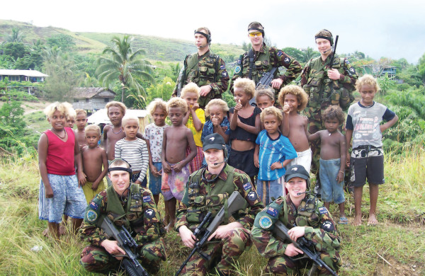 6 NZ Army soldiers smiling in uniform. They are surrounded by 16 young children on a grassy hill