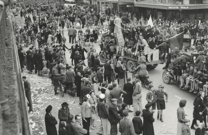 A parade of hundreds of people celebrating in the street. There are people piled onto trucks, with flags and banners.